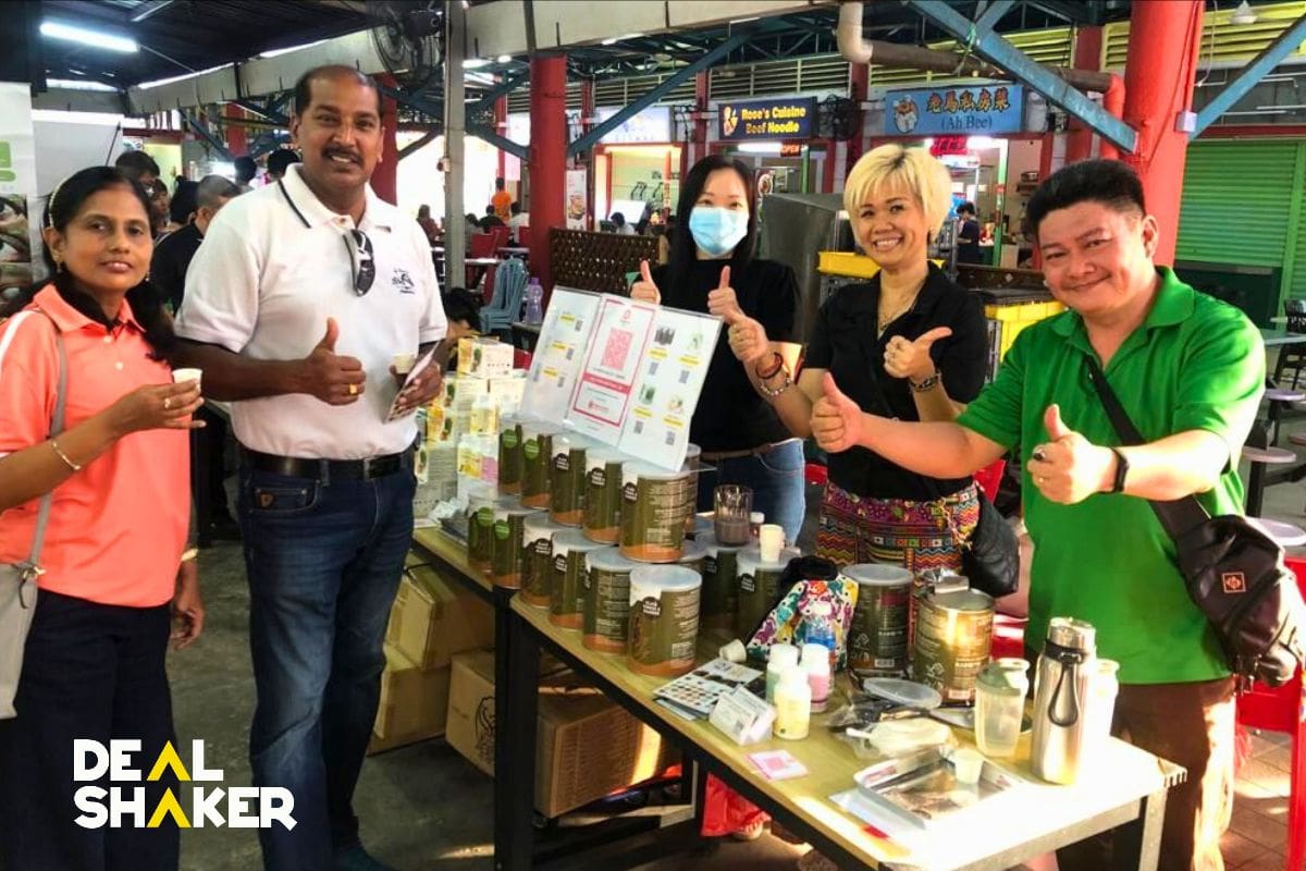 Un grup de cinc persones estan darrere d'una parada en un mercat de Malàisia, mostrant una varietat de productes amb un gest de polze cap amunt. A la cantonada inferior esquerra de la imatge s'escriu "DEAL SHAKER" i "Deal Shaker".