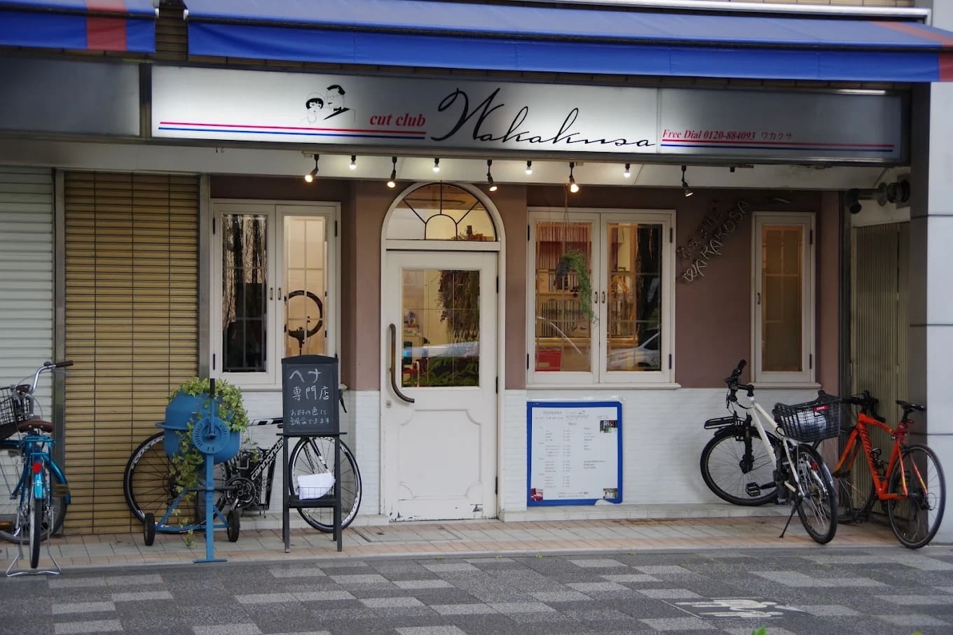 La botiga té un cartell que diu "cut club Wakana" a la part superior d'una porta doble blanca i ofereix serveis de drecera i henna. Entre les finestres i l'aparcament de bicicletes, hi ha portabicicletes blaus, plantes d'interior i menús i rètols a prop de l'entrada.