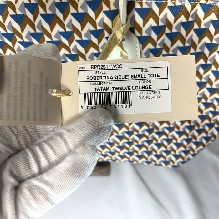 La mà enguantada sosté l'etiqueta del producte "ROBERTINA 2 (DUE) SMALL TOTE" a la part superior del teixit de tatami amb un patró geomètric en blau, marró i blanc, que és l'estil característic de la col·lecció de bosses petites de ROBERTA PIERI.