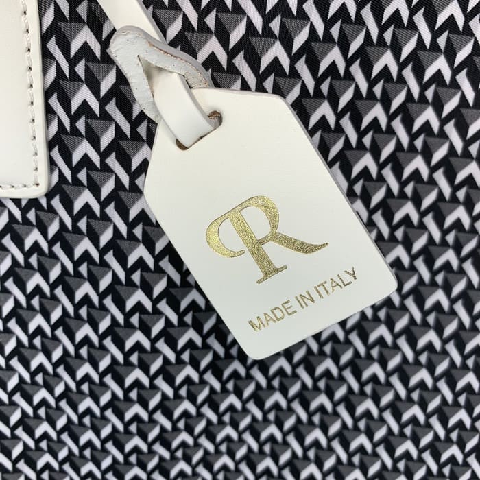 Una etiqueta ROBERTA PIERI de pell blanca amb les lletres "R" en or i les paraules "MADE IN ITALY" penja del fons d'un teixit geomètric en blanc i negre.