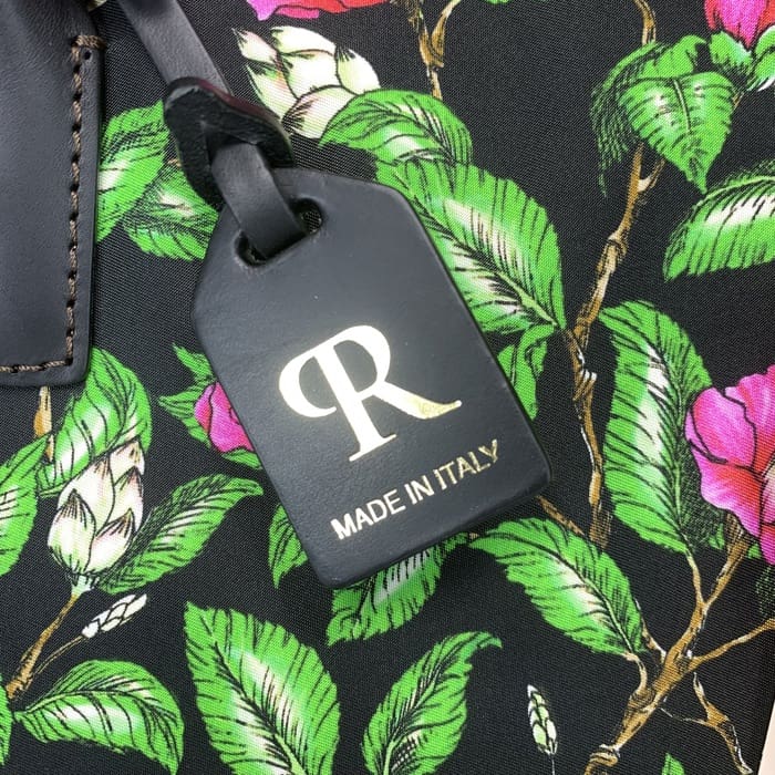 L'etiqueta d'equipatge negra de ROBERTA PIERI amb la "R" daurada i el text italià està suspesa d'un teixit floral amb fulles verdes i flors roses, complementant el mini duffel amb una elegància italiana atemporal.
