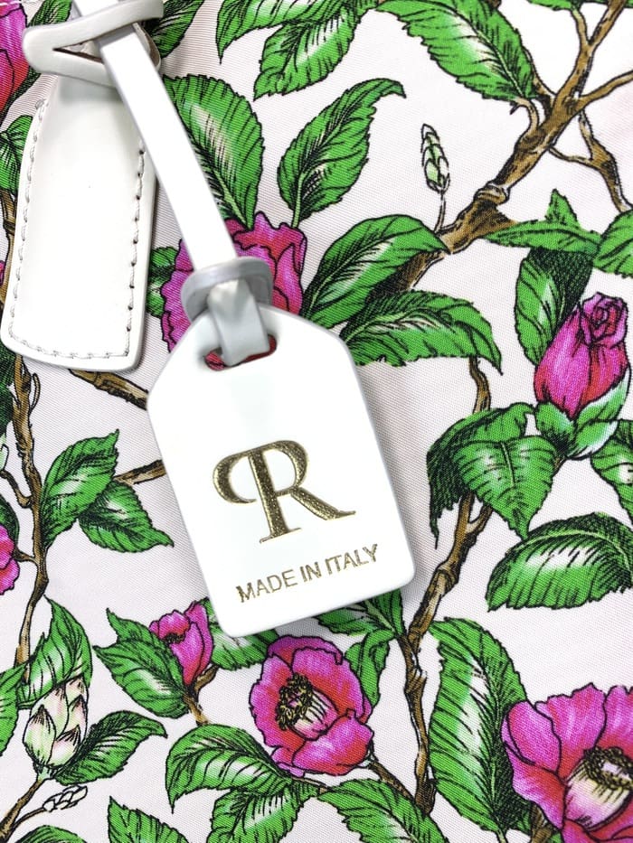 Primer pla de la mini bossa de lona ROBERTA PIERI. peduncles amb fulles verdes i flors roses; L'etiqueta blanca adjunta a la bossa de 2 vies presenta un logotip "R" daurat i les paraules "MADE IN ITALY".