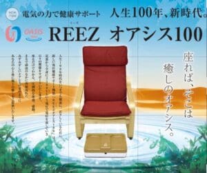 熱帯の風景を背景に、赤いクッション付きの木製椅子が置かれており、床には高陽社製のベージュ色の「REEZオアシス100 家電用電位健康サポート機器」が置かれている。画像には日本語のテキストと「OASIS 100」のブランドロゴが写っている。.