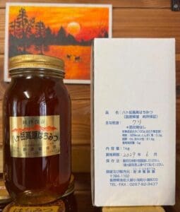 「八ヶ岳高原 国産天然はちみつ 1kg」の瓶には「純粋保証」のラベルが貼られ、ハチミツの種類として「クリ（日本の栗）」と記されており、青い日本語の文字が施された白い箱の横に置かれている。 その背後には、八ヶ岳高原の静謐な風景を彷彿とさせる、木々や山々を描いたオレンジ色の夕焼けの絵が、木製の台の上に絵のように美しい背景を演出している。.